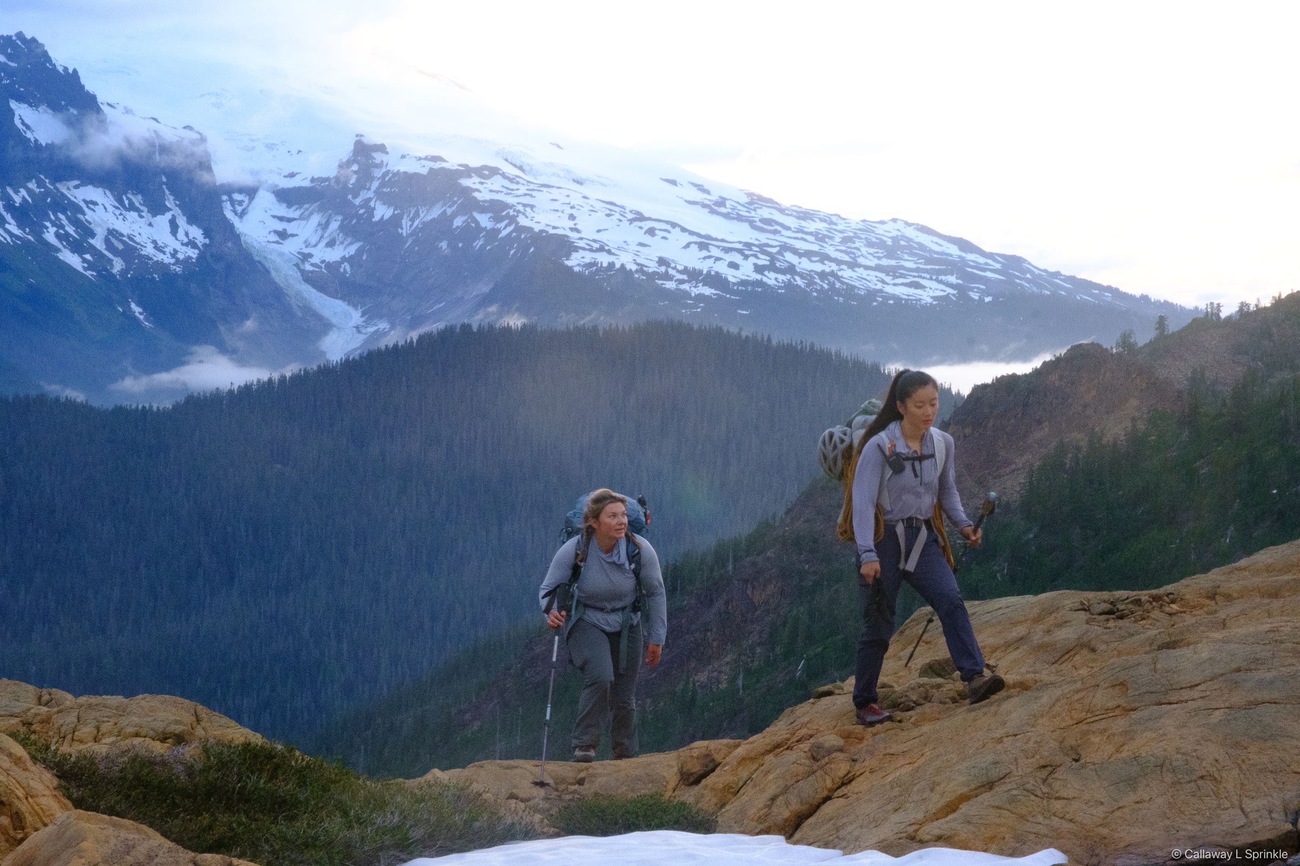 Two climbers with trekking poles crossing rust-coloured slabs, Mount Baker's glaciated massif behind