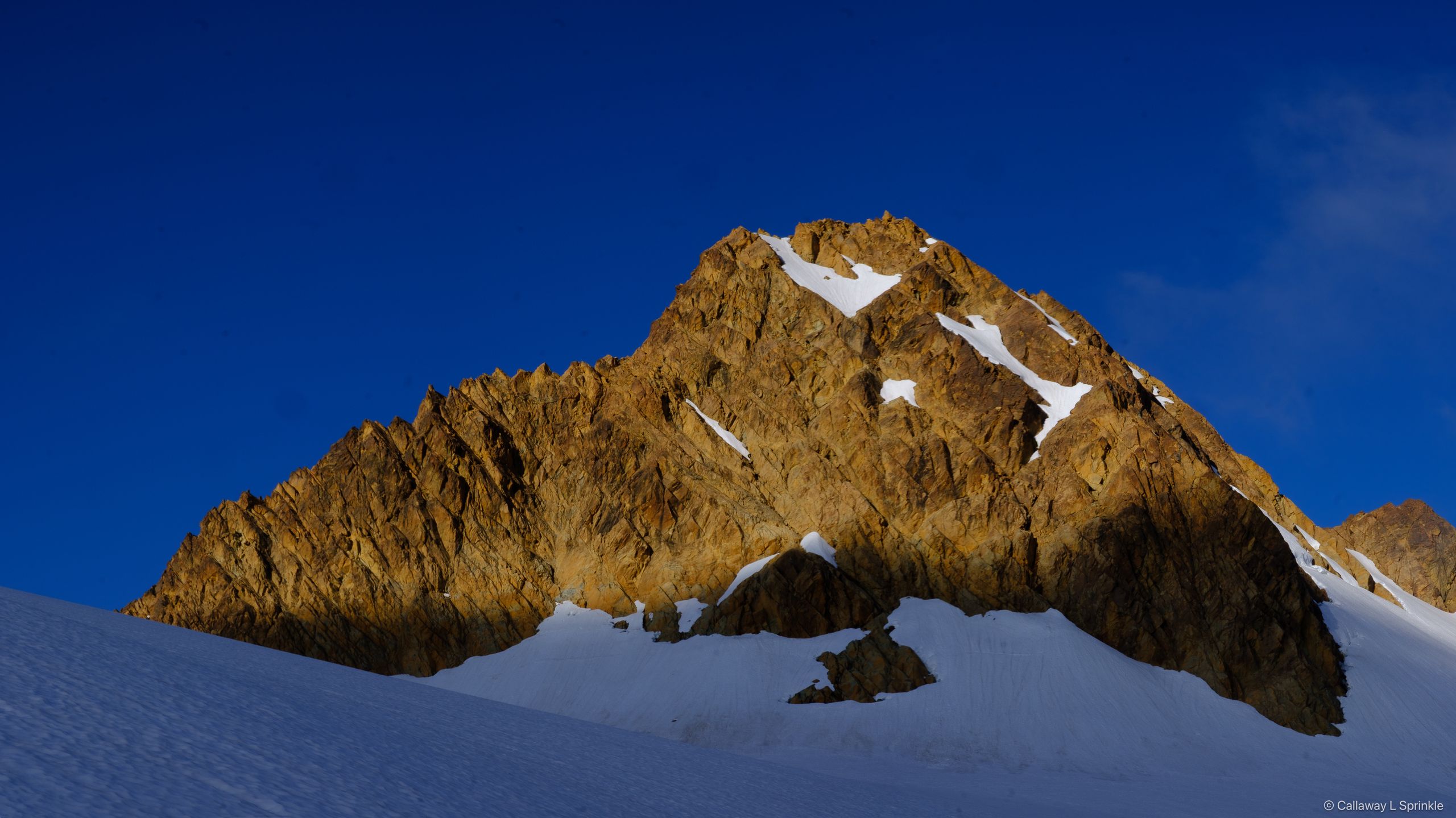 The rust-orange summit pyramid of Little Sister rising above a glacier snowfield under blue sky