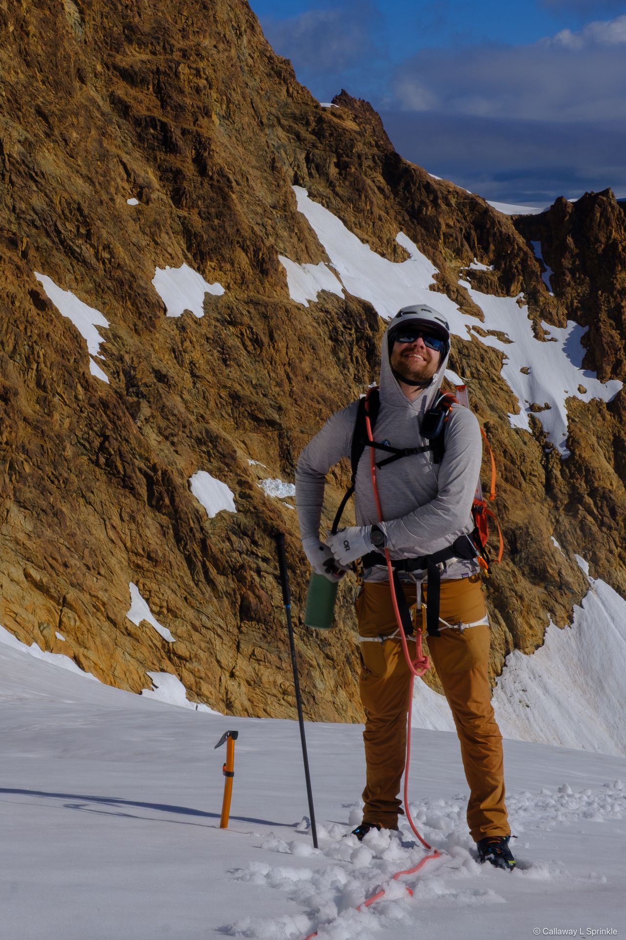 A climber standing on the glacier below the rust-orange face, ice axe planted in the snow