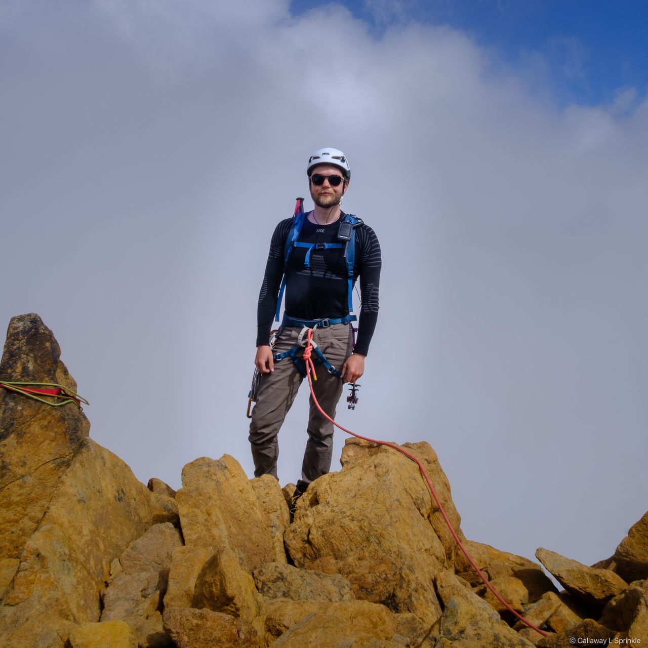 A climber standing on rust-orange summit blocks against white cloud