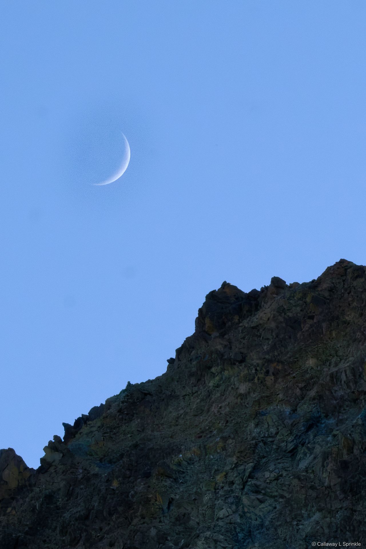 A thin crescent moon in deep blue sky above a dark ridgeline