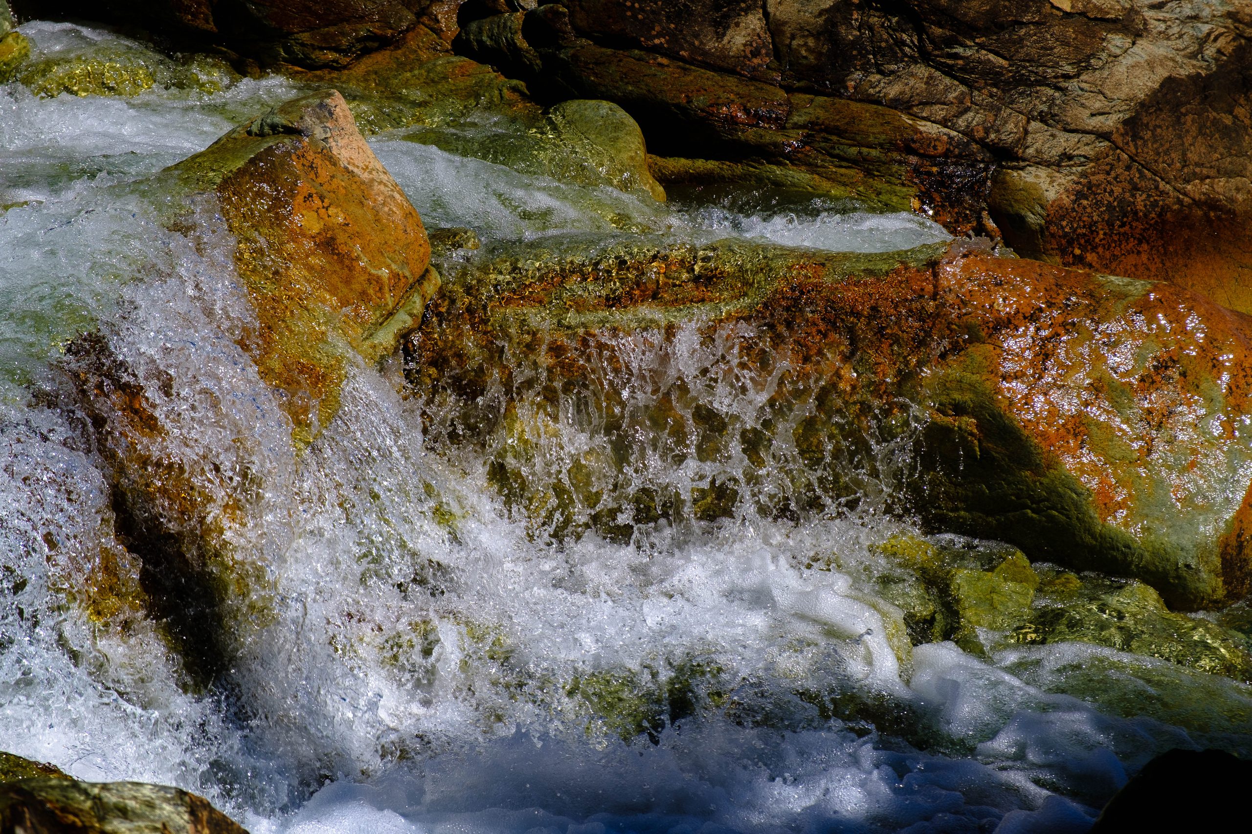 Whitewater cascading over olivine-green and rust-orange boulders in low sun, deep shadow behind.
