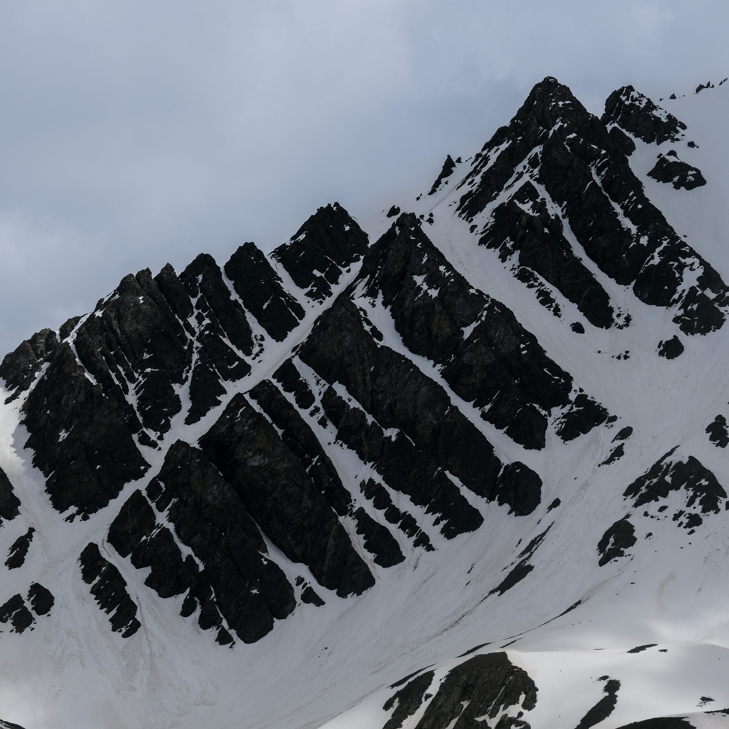 Graphic near-monochrome pattern of dark rock bands and snow on a steep ridgeline under flat grey sky.