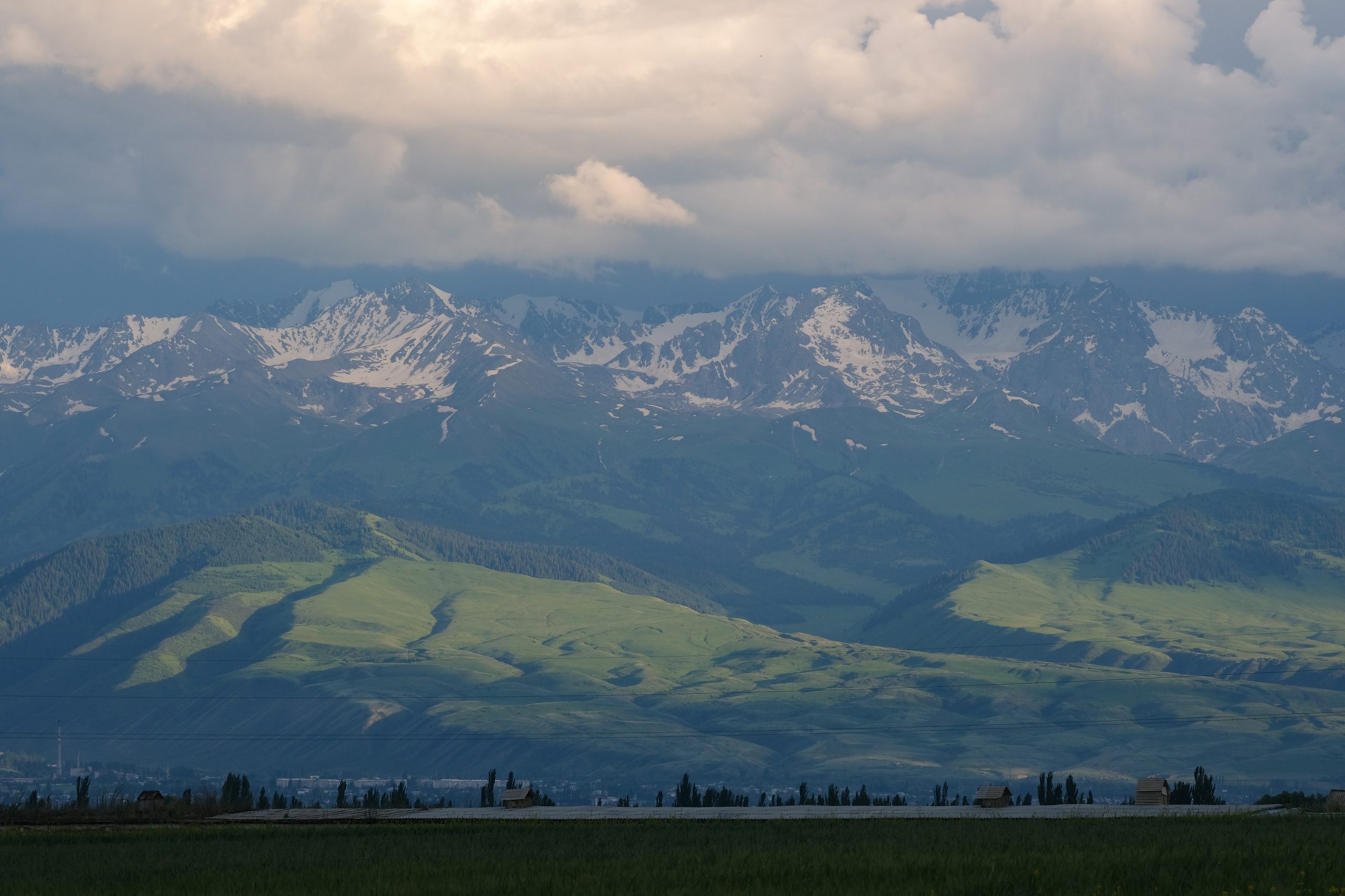 Storm light on green foothills below snow-covered peaks of the Terskey Ala-Too