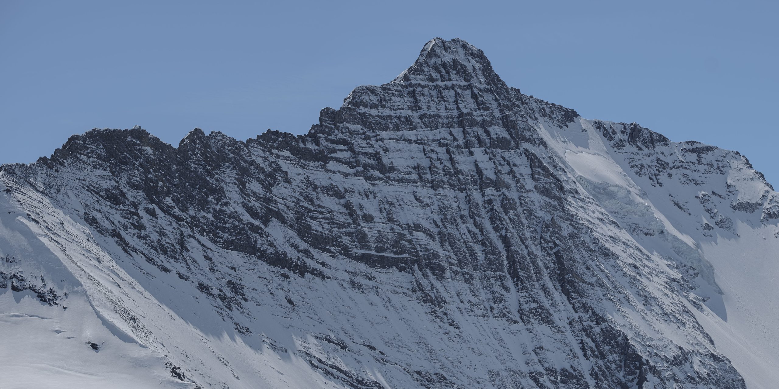 Panorama of a cold blue-grey rock pyramid streaked with snow, couloirs falling to a glacier.